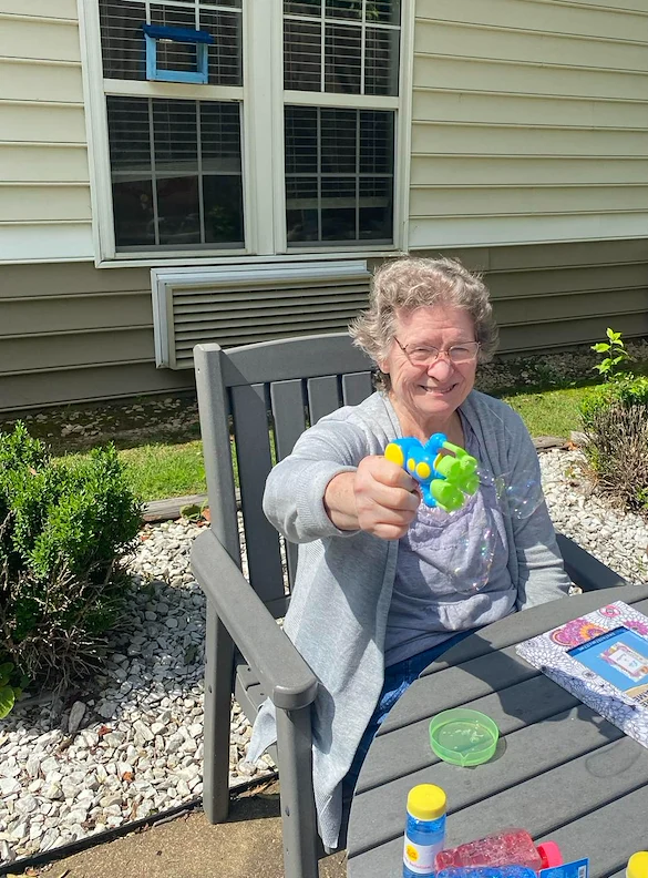 Smiling resident at Charter Senior Living of Jackson in Jackson, TN, enjoying outdoor fun with a bubble machine while sitting at a table in the sunshine, promoting happiness and wellness in assisted living and memory care.