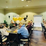 Elderly people in assisted living in Godfrey, Illinois, wearing party hats sit around a table with black and gold balloons, enjoying food and drinks amid festive New Year’s decorations. Sunlight filters through the window, creating a warm and cheerful atmosphere.