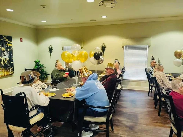Elderly people in assisted living in Godfrey, Illinois, wearing party hats sit around a table with black and gold balloons, enjoying food and drinks amid festive New Year’s decorations. Sunlight filters through the window, creating a warm and cheerful atmosphere.