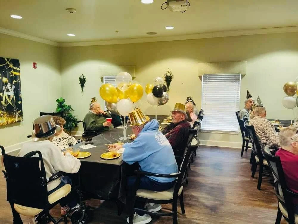 Elderly people in assisted living in Godfrey, Illinois, wearing party hats sit around a table with black and gold balloons, enjoying food and drinks amid festive New Year’s decorations. Sunlight filters through the window, creating a warm and cheerful atmosphere.