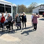 Seniors from Charter Senior Living of Jackson in Jackson, TN, smiling and waving outside a community van during an off-site trip on a sunny day, with a barn in the background, promoting social engagement and active lifestyles in assisted living and memory care.