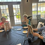 Seniors at Charter Senior Living of Hermitage in Hermitage, TN, smiling while painting individual canvases in a bright room with natural lighting, encouraging creativity and social engagement in independent living, assisted living, and memory care.