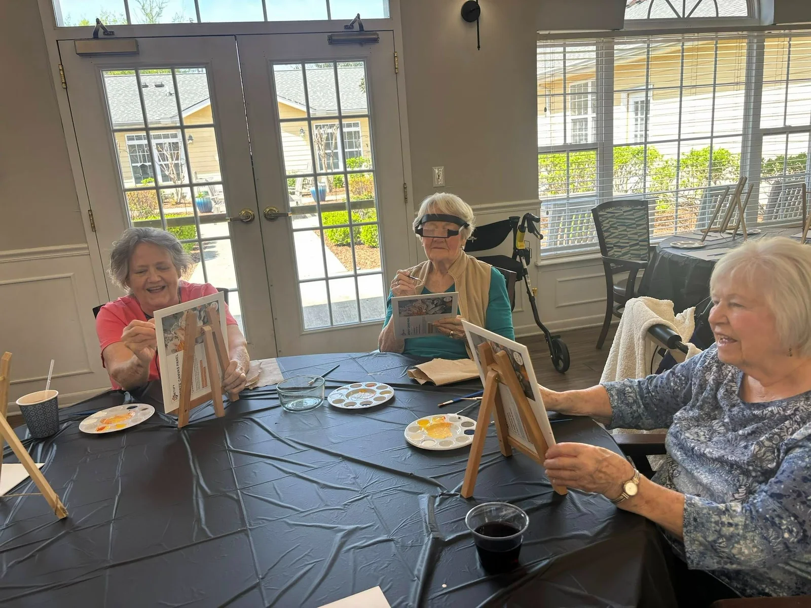 Seniors at Charter Senior Living of Hermitage in Hermitage, TN, smiling while painting individual canvases in a bright room with natural lighting, encouraging creativity and social engagement in independent living, assisted living, and memory care.