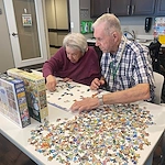 Two elderly people sit at a table covered with colorful puzzle pieces, focusing on a jigsaw puzzle. Surrounded by shelves and board games, this cozy recreation room in an assisted living community offers comfort in Godfrey, Illinois.