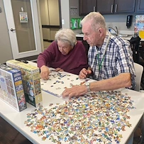Two elderly people sit at a table covered with colorful puzzle pieces, focusing on a jigsaw puzzle. Surrounded by shelves and board games, this cozy recreation room in an assisted living community offers comfort in Godfrey, Illinois.