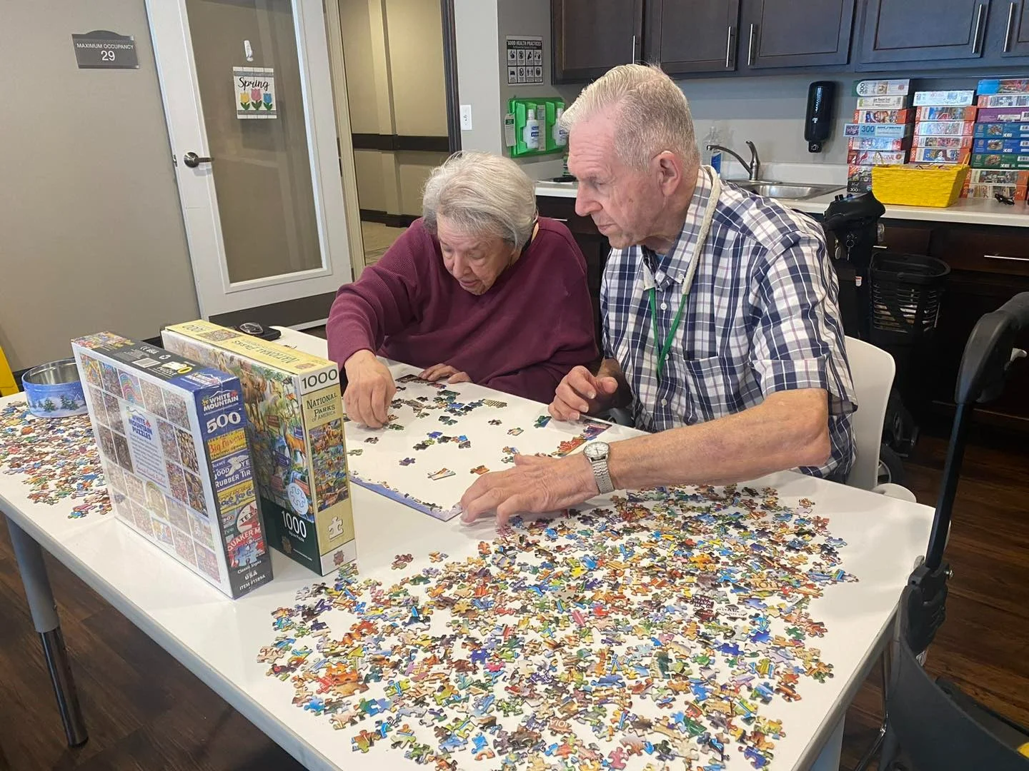 Two elderly people sit at a table covered with colorful puzzle pieces, focusing on a jigsaw puzzle. Surrounded by shelves and board games, this cozy recreation room in an assisted living community offers comfort in Godfrey, Illinois.